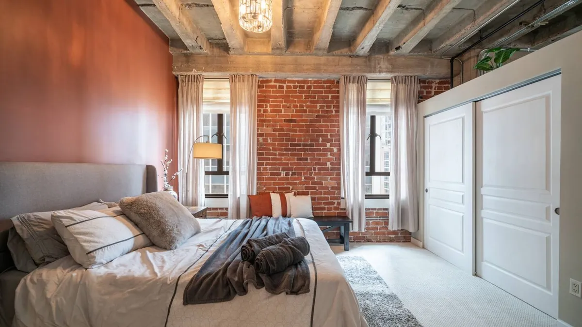 A serene bedroom featuring a plush queen bed with inviting bedding, exposed brick wall, charming light fixture, and expansive windows that flood the space with natural light. The scene exudes an inviting and relaxed mood, perfect for unwinding after a busy day.