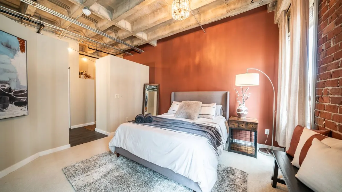 A stylish bedroom scene with a bed dressed in soft linens, a modern lamp, expressive artwork above, and an exposed brick wall. The setting is bathed in natural light, offering a comfortable and serene environment ideal for rest or a quiet morning coffee.