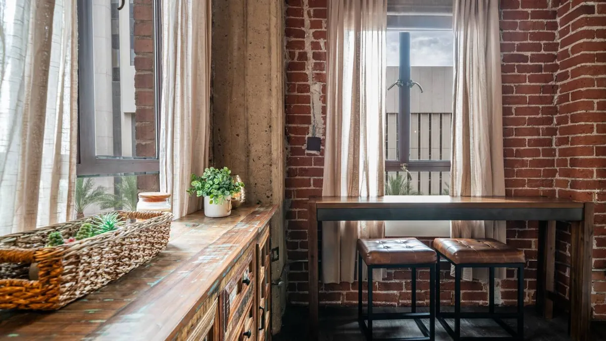 A cozy nook with a wooden countertop, exposed brick wall, window with curtains, small potted plant, and bar stools. The scene suggests a relaxed environment suitable for enjoying coffee, reading, or working on a laptop.