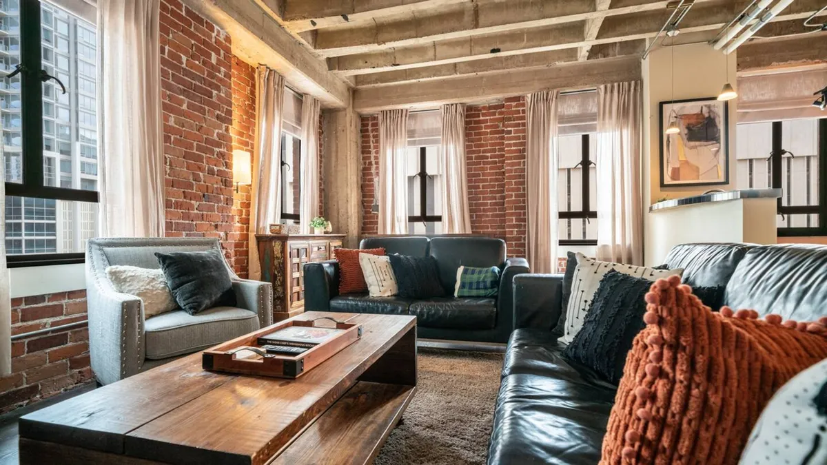 This image captures a spacious living area with exposed brick walls, comfortable leather sofas, a wooden coffee table, and large windows flooding the space with natural light. The mood is warm and welcoming, inviting guests to relax or socialize. The layout and furnishings exemplify modern industrial design while keeping the atmosphere cozy.