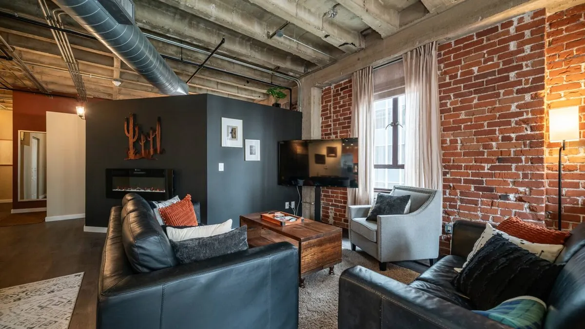The living area features exposed brick walls, comfortable seating, a wooden coffee table, a television, and eye-catching decorative wall art. The overall mood is cozy and inviting, perfect for unwinding alone or gathering with friends. Local art is an integral part of the decor, enhancing the sense of stylish hospitality.