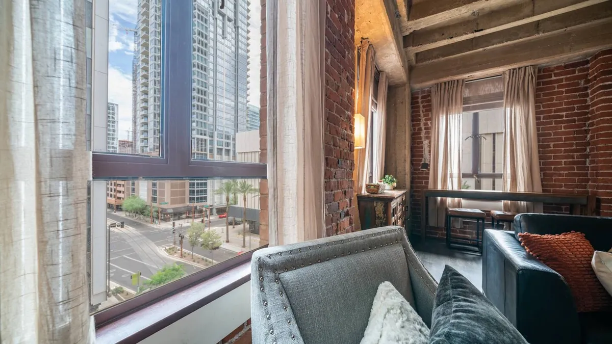 A modern seating area framed by large windows with sweeping city views, surrounded by exposed brick walls. A comfortable chair is positioned to enjoy both the urban landscape and the interior ambience, ideal for relaxing with a coffee or watching the city.