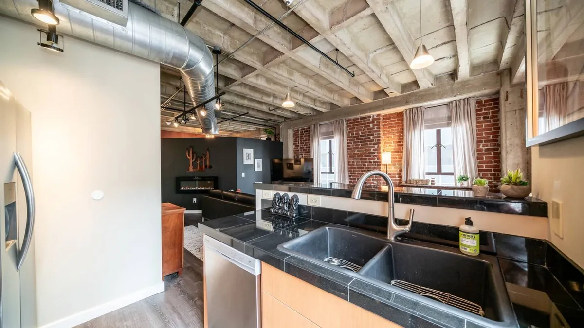 A spacious kitchen with industrial touches, exposed brick, large windows, and a kitchen island, perfect for social gatherings. The windows offer urban views, connecting indoor hospitality with the cityscape beyond.