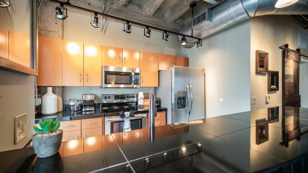 A modern kitchen featuring a large island, stainless steel appliances, wood cabinetry, and a sleek black countertop. The space is bright and inviting, with plenty of room for cooking, entertaining, or enjoying a casual breakfast.