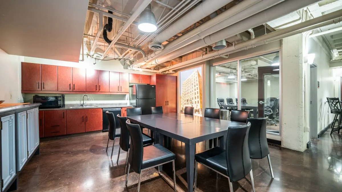 A spacious dining area with a large dining table, vibrant red cabinetry, various kitchen appliances, and a glass wall that connects the room to an adjacent space. The setting is arranged for both group meetings and casual dining, exuding a collaborative and lively atmosphere.